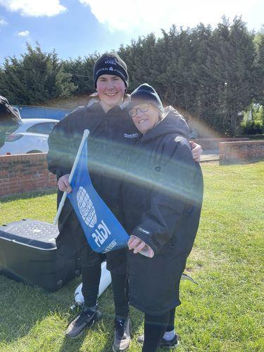 Lifestyle photo of two people in black changing robes and beanies holding a blue PADI flag beside a canoe on grass - Optimum