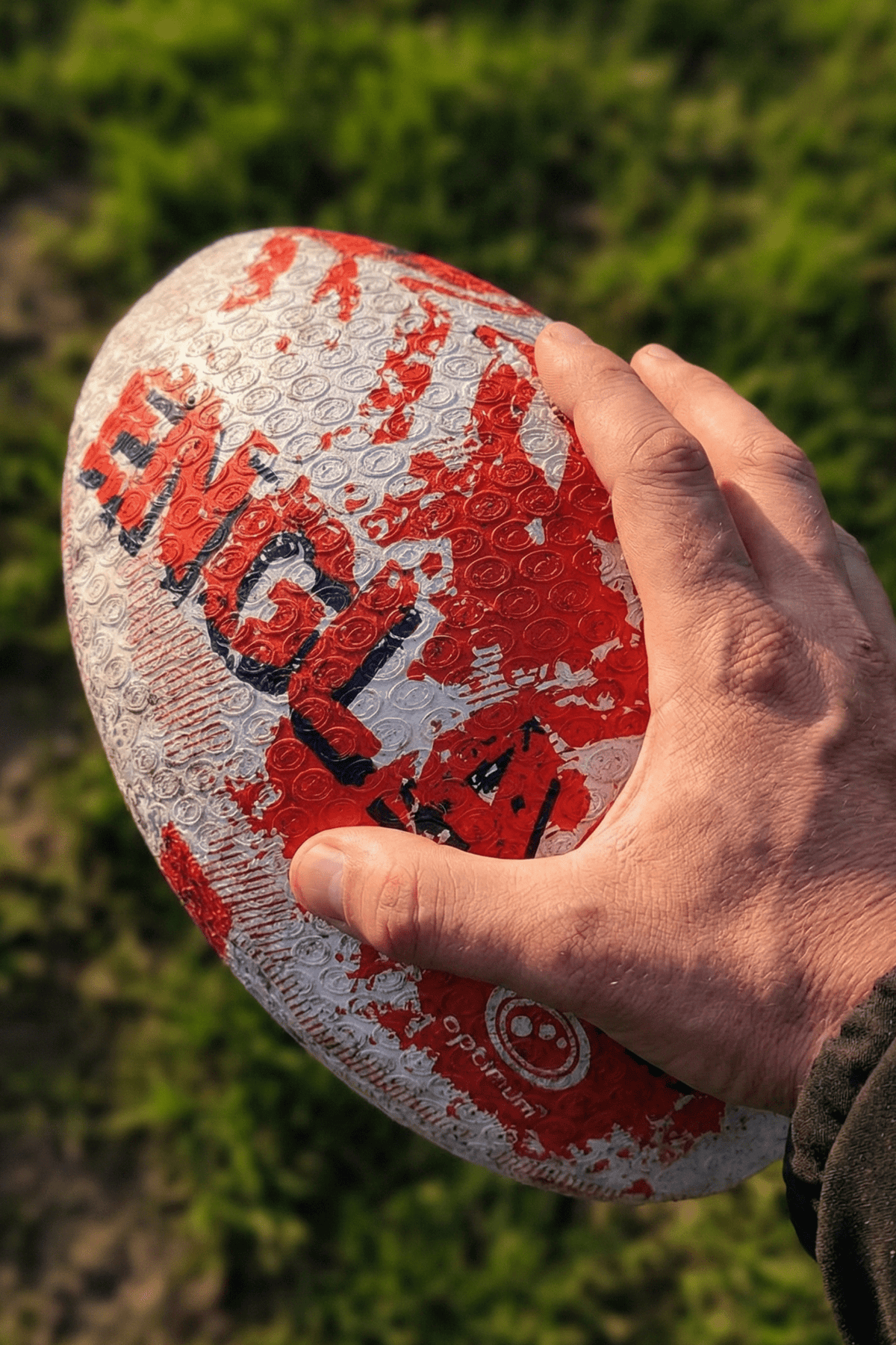 Hand holding a white and red Optimum England rugby ball with ENGLAND print above grass - Optimum