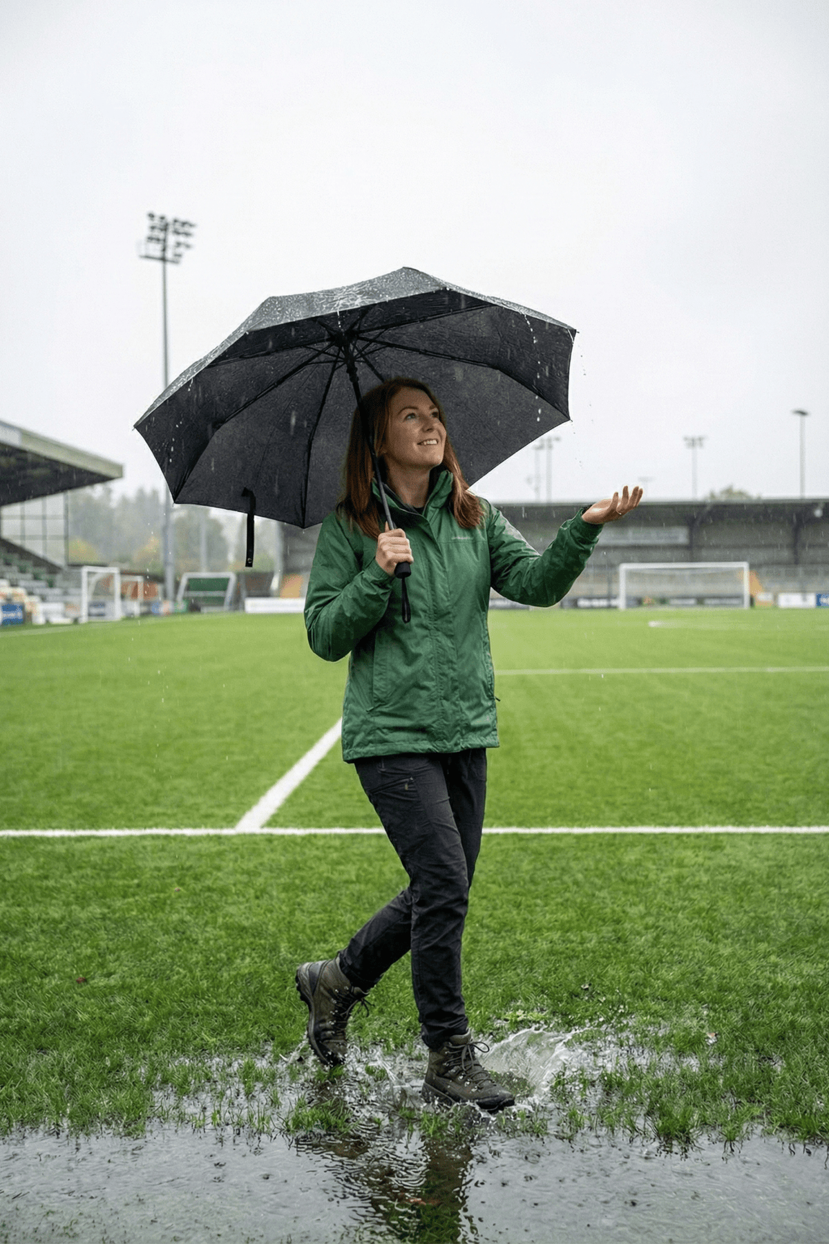 Woman holding open black Optimum umbrella on wet football pitch in rain, lifestyle shot - Optimum
